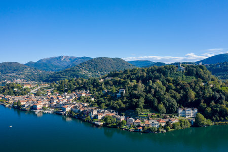 Aerial perspective of Orta San Giulio nestled along the shores of Lake Orta in northern Italy, with lush wooded hills rising behind the lakeside town. The scene features clear blue water, scattered villas, and a vivid sky creating a tranquil landscape.の写真素材