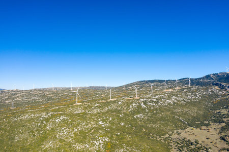 A series of wind turbines stretches across sunlit, rocky mountain slopes near Thisbe, Greece, under a deep blue sky. The landscape features scattered vegetation and rugged textures, evoking a sense of open space and clean energy.の写真素材
