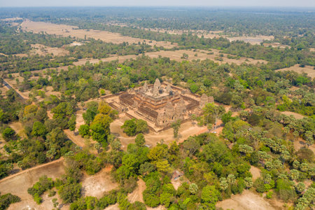 Expansive aerial perspective of the ancient Pre Rup temple complex surrounded by dry fields, scattered trees, and distant water reservoirs in the Angkor region of Cambodia. The scene captures the temples geometric layout within a patchwork of rural countryside under clear blue skies.の写真素材