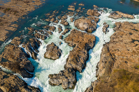 Dynamic aerial view of powerful whitewater rapids surging through rugged rocky channels at Chutes de Khone on the Mekong River in southern Laos. The dry season landscape features exposed rock formations and sparse vegetation under clear blue sky.の写真素材