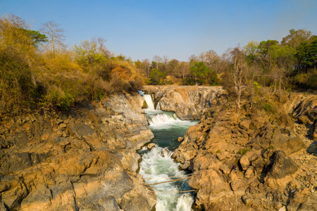 Warm sunlight illuminates the rugged rocky cascades and dry forest at Chutes de Khone on the Mekong River in southern Laos. The exposed stone and layered rapids create striking natural textures beneath a clear blue sky.の写真素材