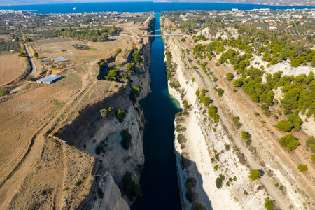 High-altitude view of the Corinth Canal cutting through steep limestone cliffs, with rural fields and scattered pine trees leading to the distant blue sea. Bright daylight highlights the dramatic landscape and clear waterway.の写真素材