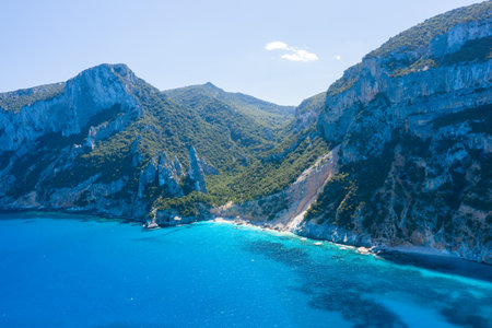 Crystal-clear turquoise water meets steep limestone cliffs and lush green slopes at Cala Goloritze, Baunei, Sardinia. Bright sunlight enhances the vivid colors and natural textures of this secluded Mediterranean cove.の写真素材