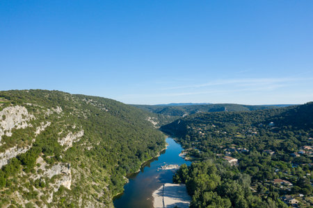 Aerial view of the winding Ardeche river cutting through steep limestone cliffs and dense green forest under a clear blue sky, with scattered rural homes dotting the landscape.の写真素材
