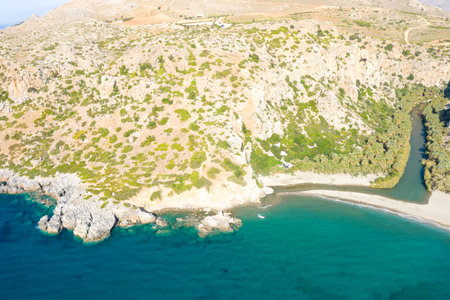 Aerial view of Preveli beach in Crete, showcasing a vivid turquoise bay, sandy shoreline, and a winding river bordered by lush greenery and rugged cliffs under bright sunlight.の写真素材