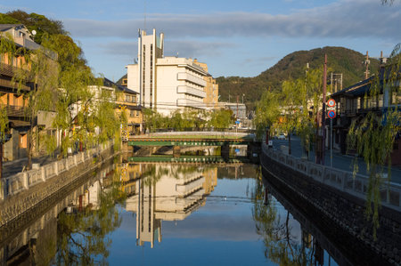 Kinosaki, Japan - November 8, 2023: A green bridge spans the central canal lined with willow trees and traditional Japanese buildings, with a modern white building and mountains visible in the background under clear daylight.のeditorial素材