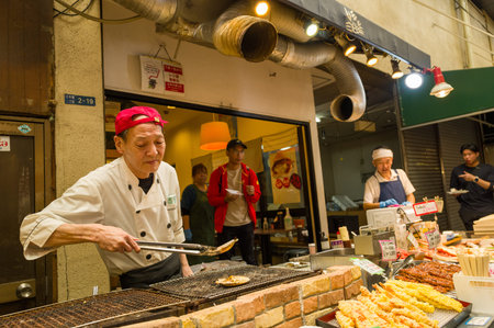 Osaka, Japan - November 7, 2023: A vendor wearing a red cap and white chef coat grills seafood on a metal grate at a market stall in Kuromon Ichiba Market, with assorted skewers and fried food displayed in the foreground and other workers and customers viのeditorial素材