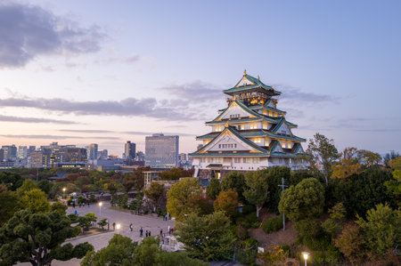 Osaka, Japan - November 7, 2023: The main tower of Osaka Castle is illuminated at dusk, with a surrounding plaza, trees, and modern city buildings visible in the background under a partly cloudy sky.のeditorial素材