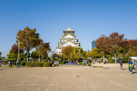 Osaka, Japan - November 7, 2023: People are seen walking and standing in an open plaza with autumn trees, with the main tower of Osaka Castle visible in the background under a clear blue sky.のeditorial素材