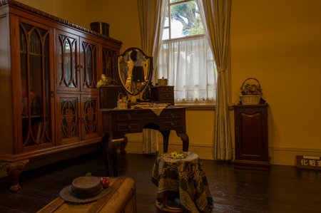 Kobe, Japan - October 30, 2023: The interior of the Moegi House is shown with a vintage wooden dressing table with a mirror, a large cabinet, a small table with a cloth, a hat and suitcase, a basket with flowers, and windows with lace curtains.のeditorial素材