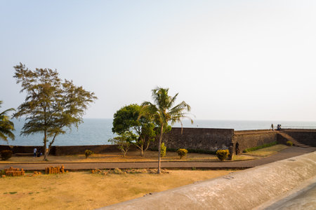 Kannur, India - March 13, 2023: The stone ramparts of Fort Kannur are visible overlooking the Arabian Sea, with several trees, a palm tree, and a dry grassy area in the foreground, and a few people standing on the fort wall under a clear sky.のeditorial素材