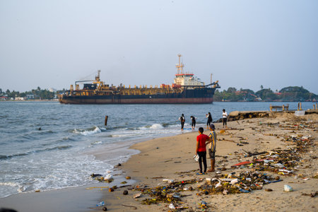 Fort Kochi, India - March 16, 2023: Several people are standing and walking along a sandy beach scattered with debris and litter, while a large cargo ship is anchored offshore in the background.のeditorial素材