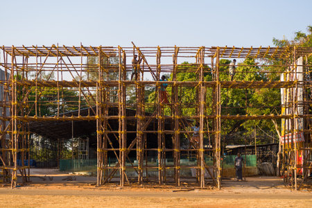 Thrissur, India - March 12, 2023: Several workers are seen assembling bamboo scaffolding for a large temporary structure on an open field, with trees and clear sky in the background.のeditorial素材