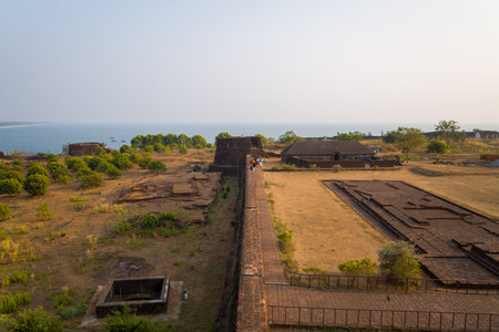 Bekal, India - March 14, 2023: An elevated view shows the laterite stone rampart running through the interior of Bekal Fort, with people walking along the wall, dry grassy areas, scattered trees, ruins, and the Arabian Sea visible in the background under のeditorial素材