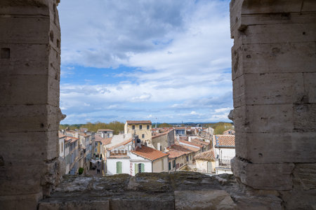 Arles, France - April 1, 2023: Rooftop view of historic buildings in Arles is seen through two large ancient stone arches, with tiled roofs and a cloudy sky in the background.のeditorial素材
