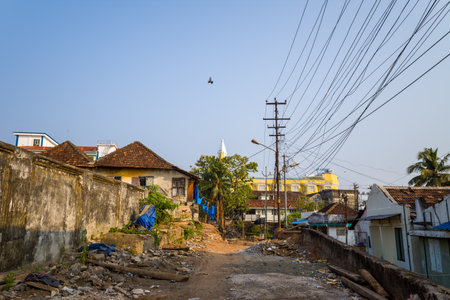 Fort Kochi, India - March 16, 2023: A residential street with scattered debris and rubble is shown in Fort Kochi, with old houses featuring tiled roofs, overhead power lines, utility poles, and a bird flying in the sky.のeditorial素材