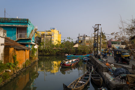 Fort Kochi, India - March 16, 2023: A canal in central Fort Kochi is lined with small wooden boats, colorful buildings, electrical poles, and auto rickshaws parked along the street, with people and a dog visible near the waters edge.のeditorial素材
