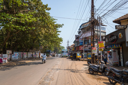Kannur, India - March 13, 2023: A main street in Kannur shows retail shops with signage in English and Malayalam, parked motorcycles, auto rickshaws, a scooter rider, pedestrians, utility poles with overhead wires, and trees lining the road under clear daのeditorial素材