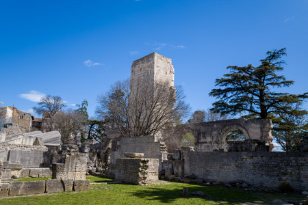 Arles, France - April 1, 2023: A large rectangular Roman stone tower stands among ancient ruins and stone arches at the site of the Ancient Theatre of Arles, with trees and a clear blue sky in the background.のeditorial素材