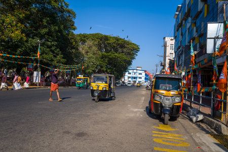 Thrissur, India - March 12, 2023: A main street in Thrissur is shown with black and yellow auto rickshaws, orange and green political party flags and banners, pedestrians walking along the sidewalk, and a commercial building under clear daylight.のeditorial素材