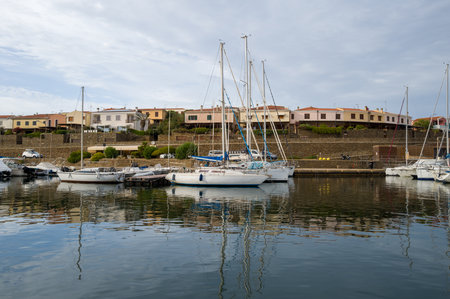 Stintino, Italy - September 13, 2023: Sailboats and motorboats are moored along the quay in the port of Stintino, with residential buildings and a stone wall visible in the background under a cloudy sky.のeditorial素材