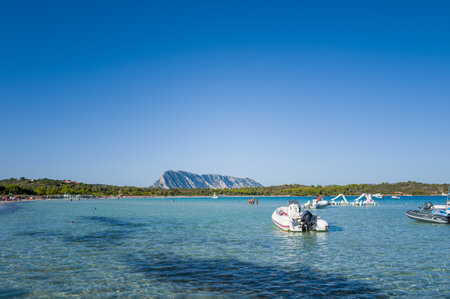 San Teodoro, Italy - September 26, 2023: Inflatable boats float on clear shallow water near Spiaggia Lu Impostu beach, with people swimming and a mountain visible in the background under a clear blue sky.のeditorial素材