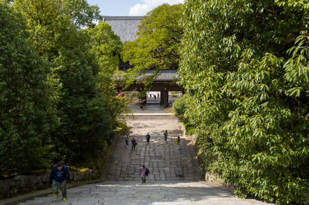 Kyoto, Japan - October 16, 2023: Several people are seen ascending a wide stone staircase lined with dense green trees, leading towards the large wooden Sanmon Gate of Chion-in Temple under a partly cloudy sky.のeditorial素材