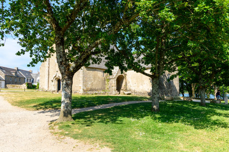 Etel, France - May 31, 2023: A stone chapel is partially visible behind several leafy trees on Ile de Saint-Cado, with a dirt path and grassy area in the foreground and traditional houses in the background under a clear sky.のeditorial素材