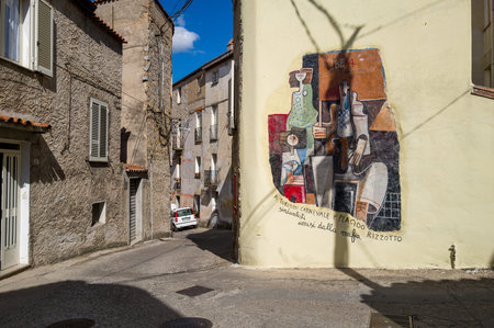 Orgosolo, Italy - September 25, 2023: A mural featuring stylized human figures and an anti-mafia dedication is painted on a yellow wall at the intersection of narrow streets, with residential stone buildings and a parked car visible.のeditorial素材