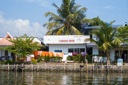 Alappuzha, India - March 6, 2023: A building with a sign reading Ayurvedic Centre is seen on the waterfront, with laundry hanging outside, palm trees, and people visible near the entrance under a clear sky.のeditorial素材