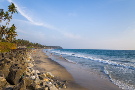 Varkala, India - March 2, 2023: People are seen walking and standing on the sandy shore of Odayam Beach, with waves reaching the coastline, large rocks in the foreground, and palm trees lining the edge of the beach under a clear sky.のeditorial素材
