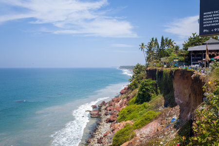 Varkala, India - March 3, 2023: The image shows a coastal cliff with the Arabian Sea below, red rocks along the shoreline, and several cafes and buildings situated at the edge of the cliff with palm trees and signage visible.のeditorial素材