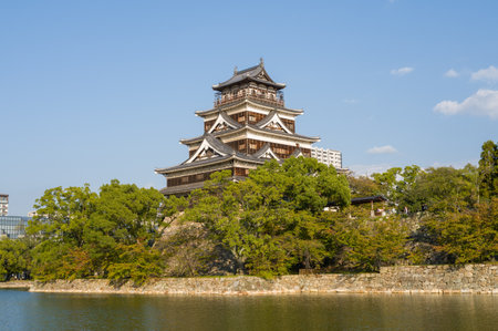 Hiroshima, Japan - November 4, 2024: The image shows Hiroshima Castle, a multi-story traditional Japanese castle structure, surrounded by trees and a stone wall, with its moat in the foreground and modern buildings visible in the background.のeditorial素材