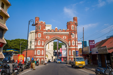 Trivandrum, India - February 28, 2023: The red brick East Fort gate stands at the center of a city street, with commercial buildings, shops, parked motorcycles, a yellow truck, cars, pedestrians, and visible signage under a clear blue sky.のeditorial素材