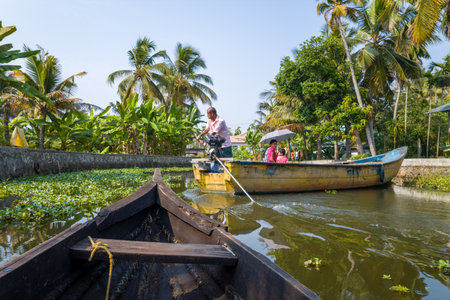 Alappuzha, India - March 7, 2023: A man operates a motorized wooden boat carrying passengers along a narrow canal lined with palm trees and tropical vegetation, with another wooden boat visible in the foreground.のeditorial素材
