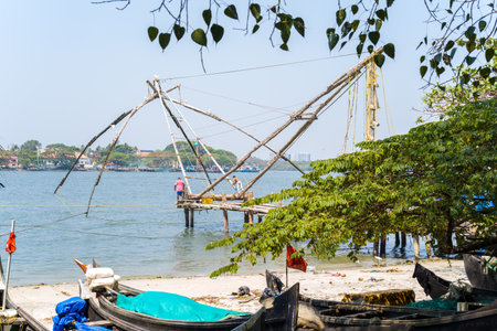 Fort Kochi, India - February 27, 2023: Two fishermen operate a large Chinese fishing net on a wooden platform extending over the water, with boats and trees visible in the foreground and buildings across the river in the background.のeditorial素材