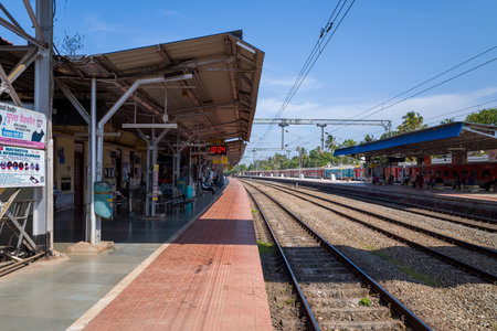 Varkala, India - March 2, 2023: Platforms and multiple railway tracks are visible at Varkala railway station, with passengers sitting and waiting under covered areas, overhead electric wires, signage, and a digital clock displaying the time.のeditorial素材