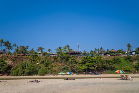 Varkala, India - March 3, 2023: People are lying on the sandy beach with colorful umbrellas and sun loungers, while cafes and palm trees line the top of the green cliff under a clear blue sky.のeditorial素材