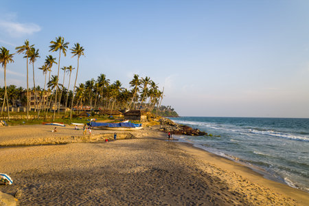 Varkala, India - March 2, 2023: People are visible on the sandy shore of Odayam Beach, with fishing boats covered by tarps, palm trees, buildings, and waves along the coastline under a clear sky.のeditorial素材