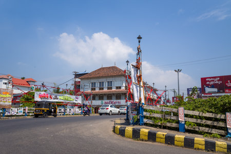 Alappuzha, India - March 7, 2023: A city center intersection features a traditional tiled-roof building, auto rickshaw, scooter, cars, banners, lamp posts, billboards, and visible signage under a clear sky.のeditorial素材