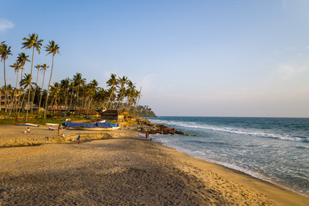 Varkala, India - March 2, 2023: People are seen on the sandy shore of Odayam Beach, with fishing boats covered by tarps, palm trees, and waves reaching the coastline under a clear sky.のeditorial素材