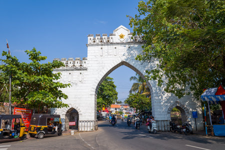 Trivandrum, India - February 28, 2023: The historic West Fort Gate, a white stone archway, stands over a busy street with auto rickshaws, motorcycles, and pedestrians passing beneath, surrounded by trees and signage.のeditorial素材