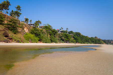 Varkala, India - March 3, 2023: A sandy beach with a shallow water pool in the foreground, dense green vegetation on the cliff, and several cafes and buildings situated at the top of the cliff under a clear blue sky.のeditorial素材