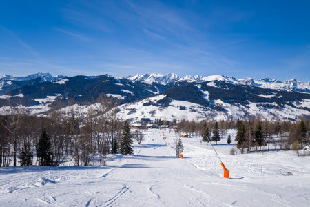 Megeve, France - January 30, 2023: A snow-covered ski slope with an orange drag lift and scattered pine trees is seen with mountain ranges in the background under a clear blue sky.のeditorial素材