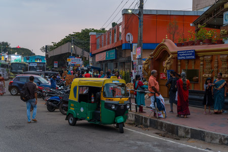 Trivandrum, India - February 28, 2023: People walk and gather near a temple chappal counter, with an auto rickshaw, motorcycles, cars, and buses visible along a busy urban street lined with commercial buildings and signage.のeditorial素材