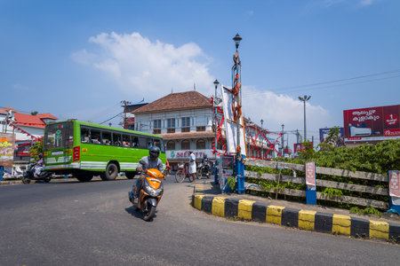 Alappuzha, India - March 7, 2023: A busy intersection features a green public bus, a person riding a scooter, and a traditional tiled-roof building, with billboards, signage, and street infrastructure visible under a clear sky.のeditorial素材