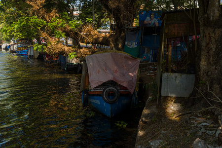 Alappuzha, India - March 6, 2023: A covered boat is moored along the shaded bank of a canal, with large trees, commercial signage, and other boats visible in the background.のeditorial素材