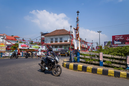 Alappuzha, India - March 7, 2023: An intersection in the city center shows people riding motorcycles and bicycles, an auto rickshaw, banners, and a traditional tiled-roof building, with signage, lamp posts, and power lines visible under a clear sky.のeditorial素材