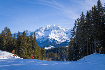 Megeve, France - February 3, 2023: A snow-covered ski slope is bordered by pine trees with a panoramic view of distant alpine mountains under a clear blue sky.のeditorial素材