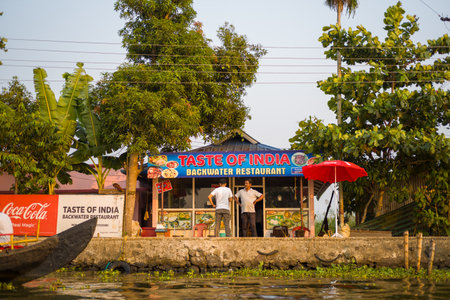 Alappuzha, India - March 7, 2023: Two men stand outside the Taste of India Backwater Restaurant, which features a prominent sign, Coca-Cola branding, a red umbrella, and is surrounded by trees along the waters edge.のeditorial素材